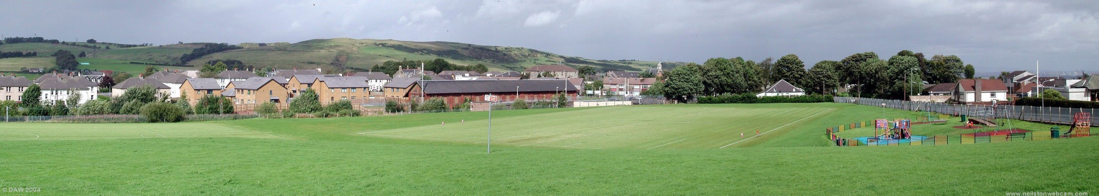 Neilston Playing fields, August 2004
The fereneze hills can be seen in the background, in the centre is the old goods shed at the railway station, now surrounded by a new housing development.  On the extreme right is the flagpole at the Neilston Bowling Club, the towerblocks of Glasgow can be seen in the distance above the club house.
