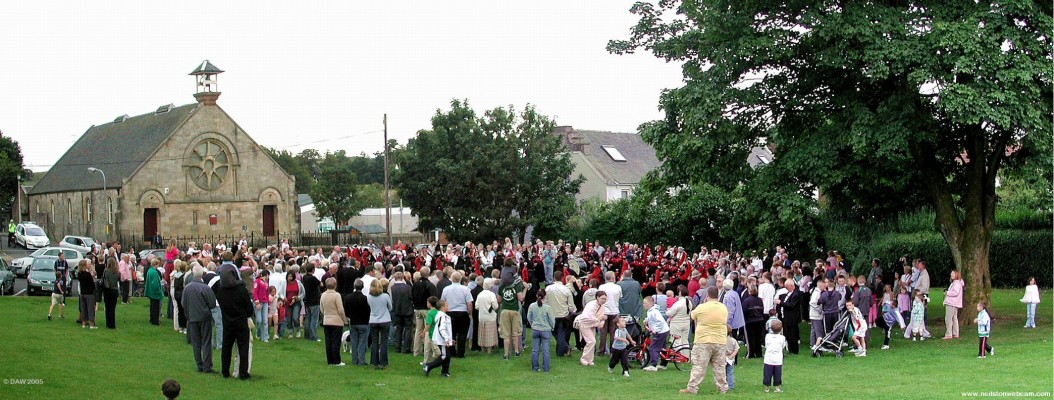 Pipe bands playing in Pig Square during the Neilston Live! event in 2005
The Royal Pipe Band of the Deputation of Ourense is in the centre of the crowd.  Neilston Parish Church Halls are in the background.
